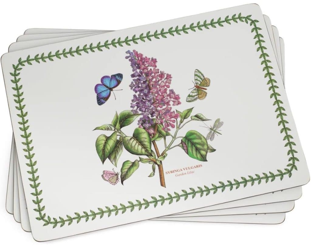 A stack of easy corkboard placemats featuring a botanical illustration of a lilac branch with green leaves, purple flowers, butterflies, and bordered by a green leafy pattern.